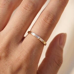 Puede incluir: Un anillo de banda dorada con tres pequeñas piedras transparentes de corte baguette rectangular, dispuestas horizontalmente en fila sobre la parte superior del anillo. El metal tiene un acabado pulido y las piedras están centradas.