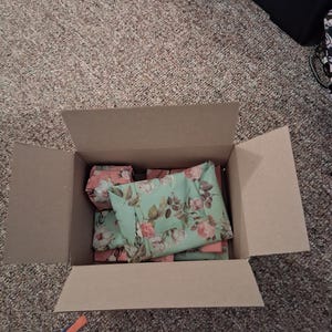 May include: An open cardboard shipping box containing several items wrapped in light mint green paper featuring a repeating pattern of pink roses, green leaves, and brown stems. The items are neatly packed inside the box, filling the interior space.