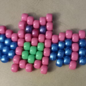 May include: A beaded bat figure in pink, blue, green, and purple. The bat is made of small, round beads, with pink beads forming the body and wings, blue beads on the wings, and green beads in the center.
