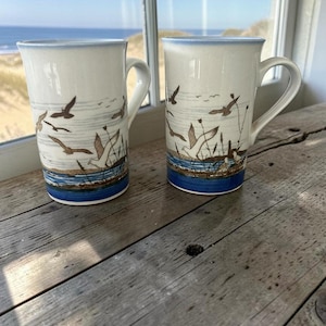 May include: Two tall ceramic mugs featuring a speckled off-white base with a coastal scene. The design includes brown flying seagulls, tall grasses, and a horizontal band of blue water at the base. Each mug has a curved handle and a light blue rim.