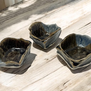 May include: Three small, dark blue and brown ceramic bowls with uneven, wavy edges. The bowls are arranged on a light wood surface.