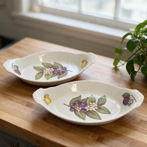 May include: Two oval ceramic baking dishes with integrated handles, featuring a white base color and a central botanical illustration of dark purple berries, green leaves, and white blossoms, with additional small fruit motifs on the sides.