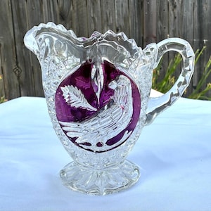 May include: A clear glass pitcher featuring a prominent circular purple glass inlay on the front. The inlay displays a detailed embossed bird and leaf design. The pitcher has a scalloped rim, a textured diamond pattern, and a sturdy handle.