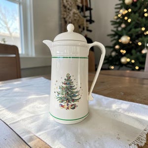 May include: A cream-colored ceramic coffee carafe with a lid and handle, featuring a central graphic of a decorated Christmas tree surrounded by small stars and wrapped gifts at the base, accented by thin green horizontal stripes near the top and bottom.