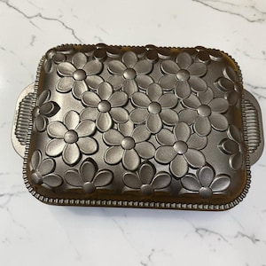 May include: A rectangular metallic baking pan with a dark bronze finish featuring a raised floral pattern across the top surface. The pan includes integrated side handles with a ribbed texture and a beaded border detail around the entire edge.