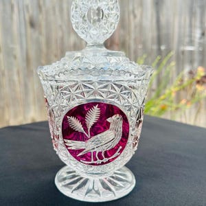 May include: A clear pressed glass candy dish with a lid featuring a faceted circular finial. The body has a geometric diamond pattern and a central circular medallion with a deep magenta background displaying a white etched bird and wheat sprig.