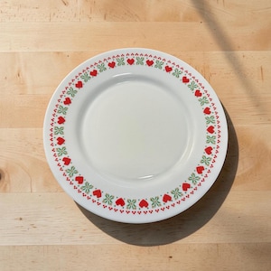 May include: A white ceramic plate with a decorative border of red hearts and green floral accents. The plate is round and sits on a light-colored wooden surface. The plate is empty.