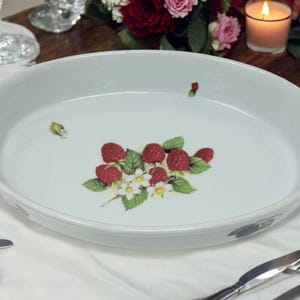 May include: White oval serving dish with a raspberry and floral design. The dish features a cluster of red raspberries with green leaves and white flowers. The rim has a subtle decorative pattern.