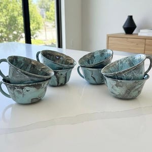 May include: A set of four ceramic cups featuring a marbled pattern in shades of teal, light blue, and dark grey. Each cup has a rounded bowl shape, a wide rim, and a single curved handle attached to the side, arranged on a white surface.