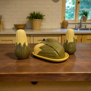 May include: A ceramic corn-shaped butter dish with a yellow base and a green lid, accompanied by two corn-shaped salt and pepper shakers. The set is displayed on a wooden surface, evoking a rustic kitchen theme.