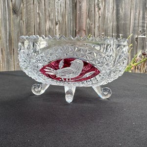 May include: A clear glass bowl with a jagged, sawtooth rim and three curved feet. The exterior features a pressed diamond pattern and a central oval panel containing a deep red inlay with an etched white bird design in the center.