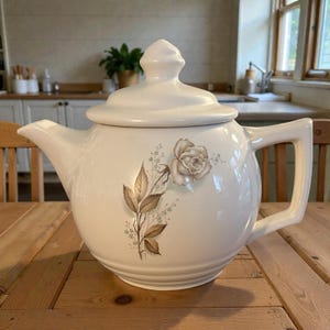 May include: Cream-colored ceramic teapot with a lid and handle. The teapot features a floral design with a rose and leaves in shades of brown and beige. The teapot is sitting on a wooden table.