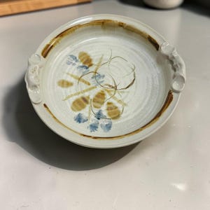 May include: A round, off-white ceramic dish with two small handles on opposite sides. The dish features a hand-painted design in the center with brown and blue floral-like shapes and circular lines, finished with a brown rim around the edge.