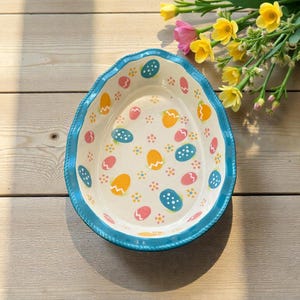 May include: An egg-shaped ceramic serving dish with a light blue rim and a white interior. The dish is decorated with colorful Easter egg designs in yellow, pink, and blue. The dish is on a wooden surface, with flowers in the background.