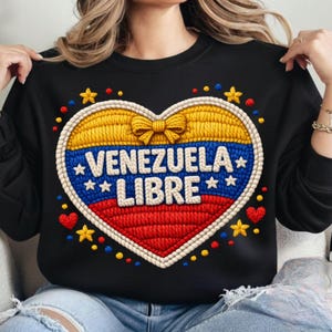 Könnte beinhalten: Schwarzes Sweatshirt mit einem herzförmigen Design mit der Aufschrift "VENEZUELA LIBRE". Das Herz ist in Gelb, Blau und Rot gehalten, mit Stern- und Herzdetails. Das Design hat ein gesticktes Aussehen.