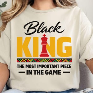 Pode incluir: Camiseta creme com as palavras "Black KING" em preto e amarelo, com uma peça de rei de xadrez vermelha. O texto "THE MOST IMPORTANT PIECE = IN THE GAME =" está abaixo da palavra "KING". Um design geométrico colorido está abaixo.