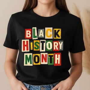 May include: Black t-shirt with the words "BLACK HISTORY MONTH" in colorful, block letter design. The letters are in red, green, yellow, and white, set against a black background. The shirt is short-sleeved.