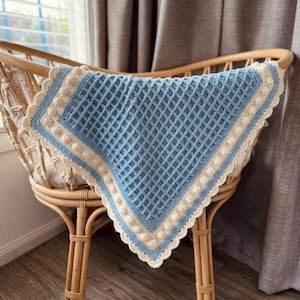 Può includere: Una coperta per neonati fatta a mano con un motivo a punto waffle azzurro, rifinita con un bordo color crema e un bordo smerlato. La coperta è esposta in una culla di vimini, che mette in risalto la sua morbida consistenza e il design.