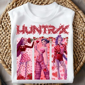 May include: White t-shirt featuring the word "HUNTRAX" in red letters with a heart design. The shirt displays three cartoon figures in pink outfits, surrounded by hearts and roses, perfect for Valentine's Day.