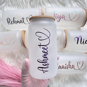 May include: Frosted glass beverage containers with bamboo lids. Each container has a name in a script font. The names are in various colors, including purple and rose gold. The containers are on a white, fluffy surface.