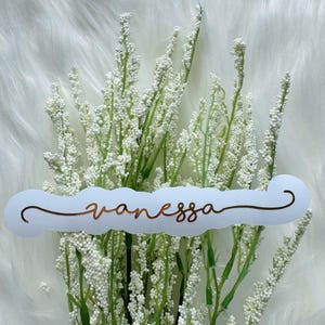 Pode incluir: Uma etiqueta de nome em forma de nuvem branca com o nome "Vanessa" em escrita cor de cobre. A etiqueta está colocada sobre um fundo de flores brancas com caules verdes, tudo sobre uma superfície branca e fofa.