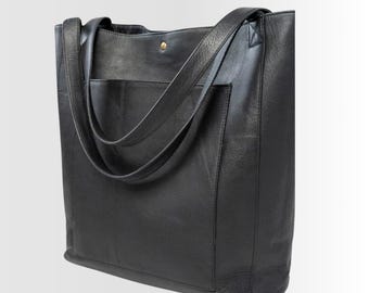 以下が含まれることがあります： 前面に2つの大きなポケットが付いた黒い革製のトートバッグ。肩掛け用の長いストラップが2本付いています。
