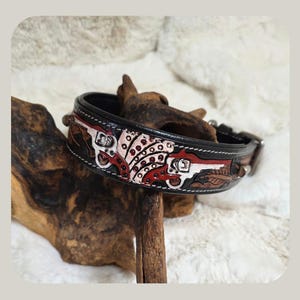 May include: Black leather dog collar with a decorative design featuring red, white, and brown accents. The collar has a detailed pattern with what appears to be a gun and other western-themed elements. The collar is secured with a metal buckle.