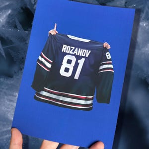 Könnte beinhalten: Blaue Karte mit einem Hockey-Trikot-Design. Das marineblaue Trikot zeigt den Namen "ROZANOV" und die Nummer "81" in Weiß. Rote und weiße Streifen zieren die Ärmel und den Saum. Die Nummer "8" ist ebenfalls sichtbar.