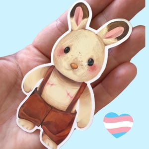 May include: A white plush bunny with brown ears and pink accents. The bunny is wearing brown overalls and has a stitched scar on its chest. The bunny is holding a small heart with the transgender pride flag colors.