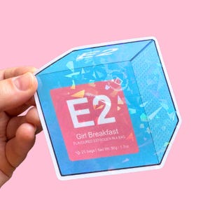 May include: A holographic sticker in the shape of a cube, held against a pink background. The sticker features the text "E2" in large white letters on a red background, with the words "Girl Breakfast" and "Flavored Estrogen in a Bag" below.