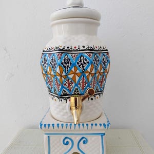 Peut inclure: Distributeur de boissons en céramique blanche avec couvercle, orné d'un motif géométrique coloré bleu, orange et noir. Il possède un robinet doré et repose sur un socle blanc assorti avec des accents bleus. Une pièce décorative pour servir des boissons.