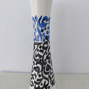 Può includere: Un vaso in ceramica bianca con base svasata e collo stretto. Il vaso è decorato con motivi calligrafici blu e neri. La sezione superiore presenta forme astratte blu, mentre la sezione inferiore mostra la calligrafia nera.
