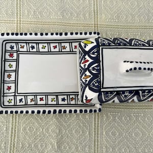 May include: A white ceramic butter dish and matching rectangular tray. The dish and tray are decorated with a black border and colourful floral accents. The lid has a small handle. The tray has a handle on one side.
