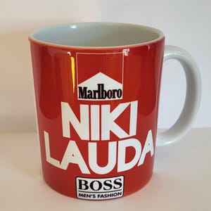 May include: Red ceramic mug with a white design featuring the words "Niki Lauda" and the Marlboro logo. The bottom of the mug has the words "Boss Men's Fashion."