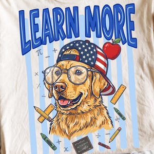 May include: A cream-colored t-shirt featuring a graphic of a golden retriever wearing glasses and a baseball cap with an American flag pattern. The text "LEARN MORE" appears in blue above the dog, surrounded by illustrations of an apple, pencils, and math symbols.