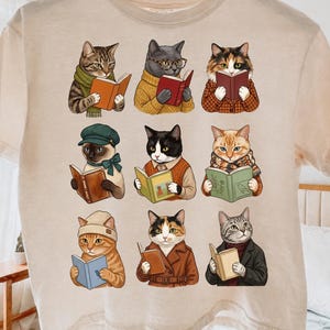 以下が含まれることがあります： ベージュのTシャツで、それぞれ異なる服を着て本を読んでいる9匹の猫のグラフィックプリントが施されています。猫は3×3のグリッド状に配置されており、タビー、三毛猫、タキシード猫など、さまざまな品種や色が描かれています。