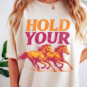 Puede incluir: Camiseta color crema con las palabras "HOLD YOUR" en naranja y magenta. Debajo del texto, dos caballos corriendo en naranja y magenta. La camiseta es de estilo casual y unisex.