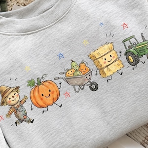 Könnte beinhalten: Ein hellgraues Sweatshirt mit Rundhalsausschnitt und einem farbenfrohen Grafikdruck, der eine lächelnde Vogelscheuche, einen großen orangefarbenen Kürbis, eine mit Kürbissen gefüllte Schubkarre, einen Heuballen und einen grünen Traktor zeigt.