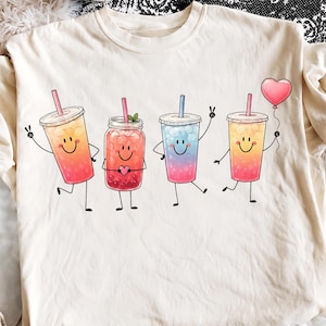 Może przedstawiać: Kremowy t-shirt z grafiką przedstawiającą cztery antropomorficzne kubki z napojami, posiadające uśmiechnięte buzie, ręce i nogi. Napoje obejmują mrożone herbaty i słoik, przy czym jeden kubek trzyma różowy balon w kształcie serca.