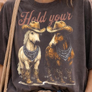 May include: Dark gray t-shirt featuring a graphic of two horses wearing cowboy hats, bandanas, and boots. The text "Hold your" is arched above the horses in a reddish-brown color. The shirt has a vintage, worn look.