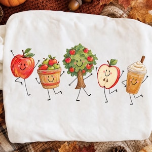 Puede incluir: Una sudadera blanca con un gráfico colorido de cinco personajes antropomórficos: una manzana roja, una cesta de madera llena de manzanas, un manzano, una manzana cortada y una bebida beige en un vaso, todos con caras sonrientes.