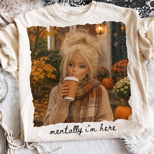 Op de afbeelding: Een cr&egrave;mekleurig t-shirt met een rechthoekige afbeelding van een persoon met blond haar in een bruine trui en sjaal, die een koffiebeker vasthoudt in een herfstsetting met pompoenen. De tekst "mentally i'm here" is in het zwart onder de afbeelding gedrukt.