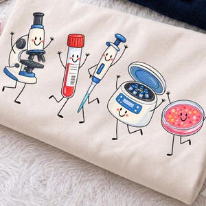 May include: A cream-coloured fabric featuring a graphic print of five animated laboratory items with smiling faces: a blue and white microscope, a red-capped test tube, a blue and white pipette, a centrifuge, and a pink petri dish with colourful dots.