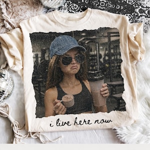 May include: Cream-colored t-shirt featuring a distressed image of a person wearing a denim cap and sunglasses, holding a protein shake and scoop. The text "i live here now" is written in a handwritten style at the bottom of the image.