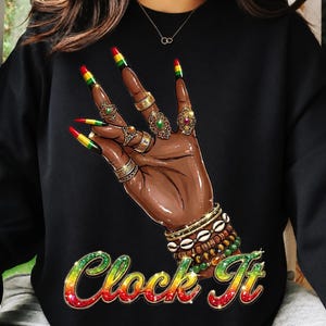 Puede incluir: Sudadera negra con un gráfico de una mano adornada con anillos, pulseras y uñas largas pintadas en rojo, amarillo y verde. El texto "Clock It" está escrito en un degradado arcoíris.