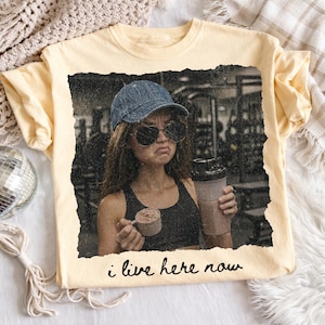 May include: A light yellow t-shirt featuring a distressed image of a person in a gym setting, holding a protein shaker and scoop. The text "i live here now" is printed at the bottom of the image.