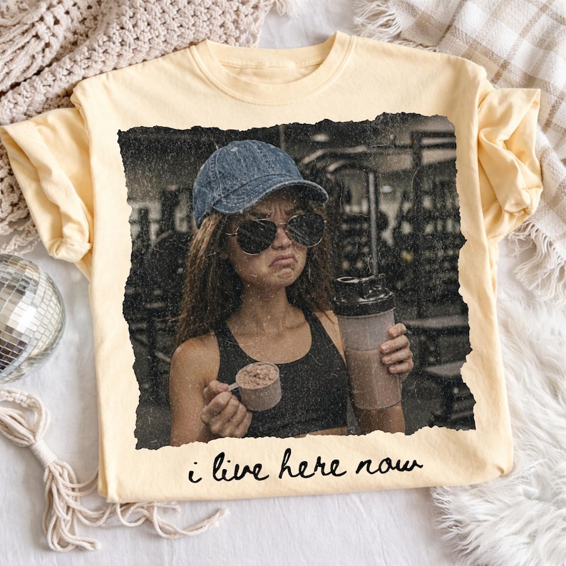 May include: A light yellow t-shirt featuring a distressed image of a person in a gym setting, holding a protein shaker and scoop. The text "i live here now" is printed at the bottom of the image.