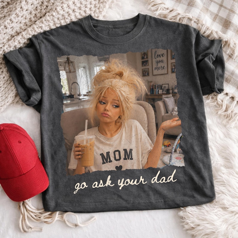 May include: Dark gray t-shirt featuring a distressed image of a woman holding a drink, with the word "MOM" and the phrase "go ask your dad". A red baseball cap is visible on the left.