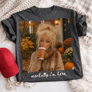 Op de afbeelding: Een donkergrijs t-shirt met een grafische print van een persoon met blond haar in een beige trui en sjaal, die een koffiebeker vasthoudt, omringd door herfstbladeren en pompoenen, met de witte tekst "mentally i'm here" aan de onderzijde.
