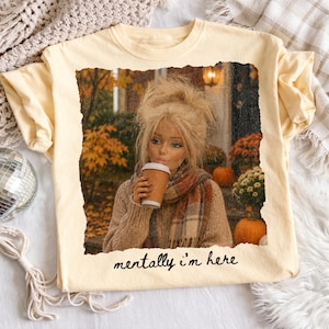 Op de afbeelding: Een lichtgeel t-shirt met een rechthoekige grafische print van een blonde pop met warrig haar, gekleed in een bruine trui en een geruite sjaal, terwijl deze een koffiebeker vasthoudt. De achtergrond toont herfstbladeren en pompoenen. De tekst luidt mentally i'm here.